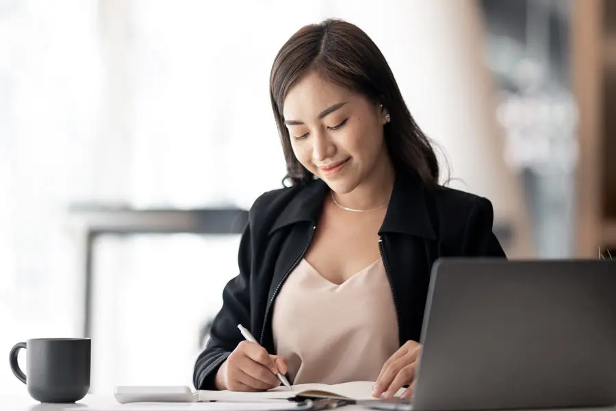 Businesswoman writing in notebook at desk with coffee.