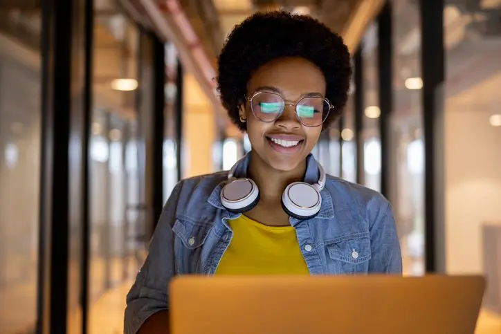 Woman with glasses and headphones smiling while using a laptop.