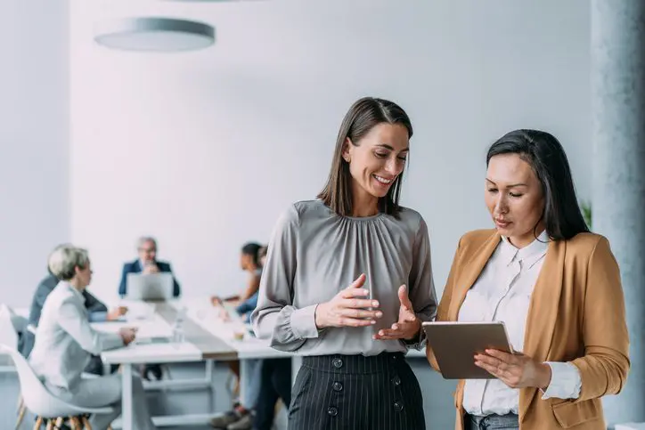 Two businesswomen reviewing a tablet with colleagues in the background.