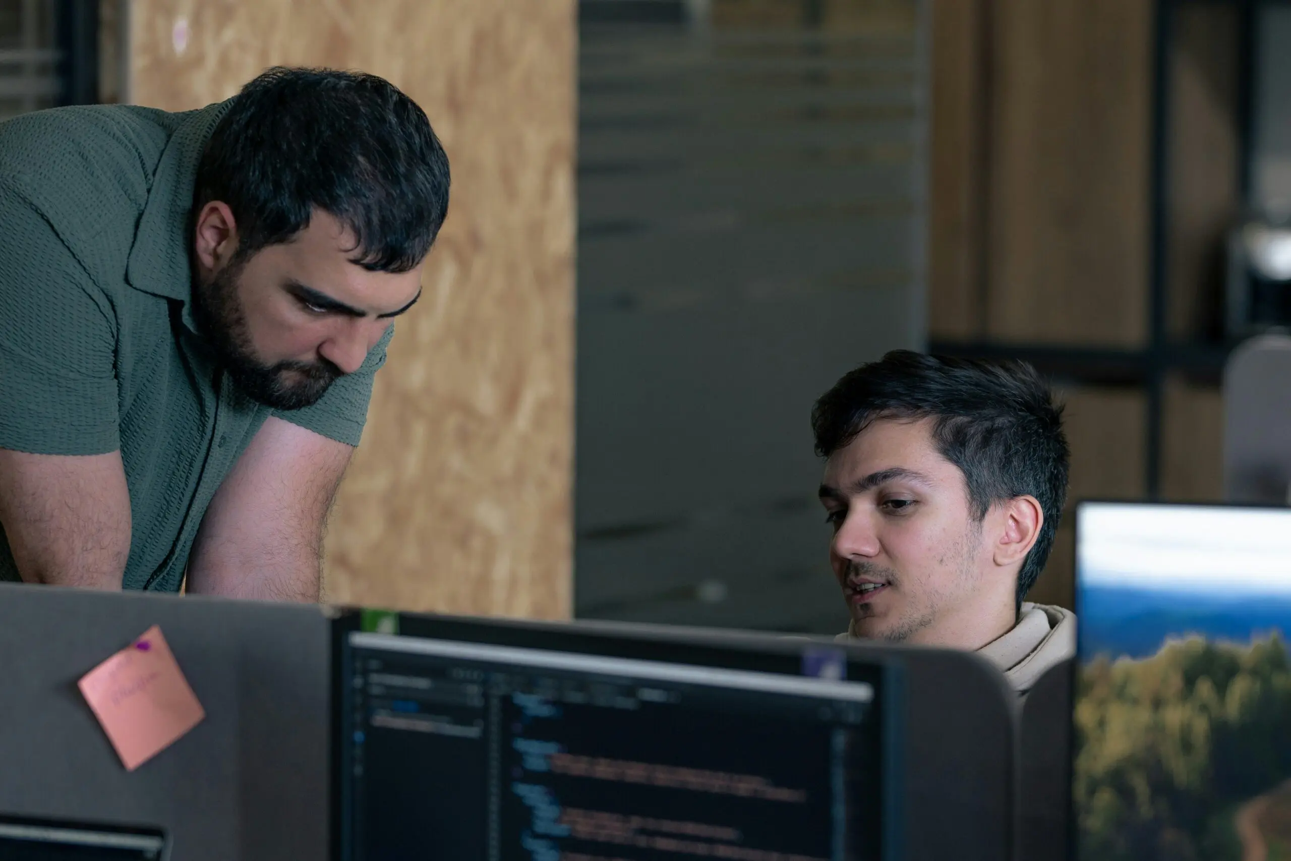 Two male developers discussing code at computer workstations in an office.
